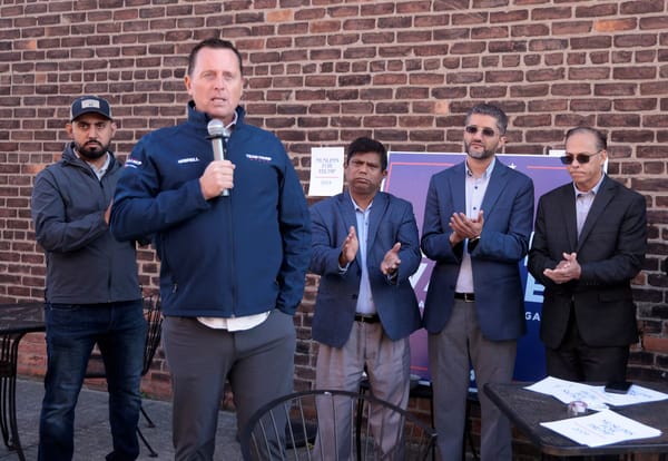 A man speaks into a microphone while four men behind him applaud.